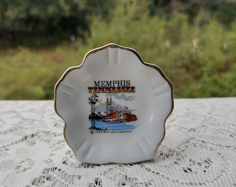 Könnte beinhalten: Eine weiße Keramikschale mit goldfarbenem Rand, die eine farbenfrohe Illustration eines Dampfschiffs auf dem Mississippi in Memphis, Tennessee, zeigt. Der Text "Memphis Tennessee" ist in roten und schwarzen Buchstaben gedruckt. Die Schale ist mit einem goldenen Rand und einem Copyright-Hinweis für "Deep South Graphics" verziert.
