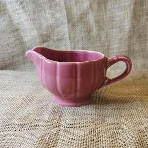 May include: A small, antique-style, mauve-colored creamer with a handle and a spout. The creamer has a ribbed, scalloped design around the body. The handle is curved, and the spout is pointed. The creamer is made of ceramic.