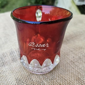 May include: A deep red glass mug featuring a clear, textured base with a scalloped design. The word Oscar is etched in white script on the front of the red glass surface. The mug has a wide, flared rim and a clear glass handle visible at the top.