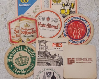 May include: A collection of nine different beer coasters, featuring various brewery logos and designs. The coasters are round and square, with a variety of colors and patterns. Some of the coasters feature images of beer mugs, while others feature text.