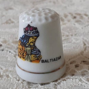 May include: A white ceramic thimble featuring a colorful illustration of a king wearing a crown and holding a small vessel. The name BALTIAZAR is printed in black capital letters next to the figure. The base has a thin gold trim line.