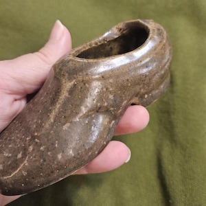 May include: A handmade ceramic shoe-shaped planter in shades of brown and tan. The open top of the shoe provides space for small plants or decorative items. The planter has a rustic, textured finish.