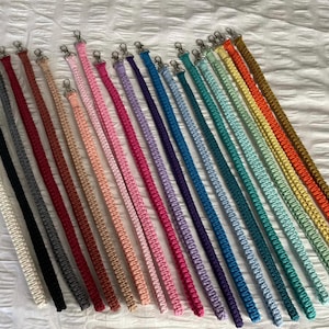 May include: A selection of macrame bag straps in a spectrum of colours, such as black, cream, grey, red, pink, purple, blue, green, yellow, and orange. Each strap features a silver-tone clasp.