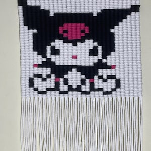 以下が含まれることがあります： ピンクのリボンとフリンジが付いた白黒のピクセルアートの壁掛け。大きな目の漫画のキャラクターのデザインです。