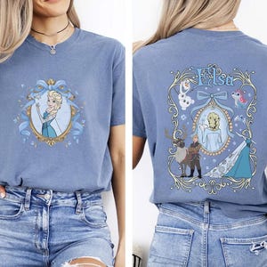 May include: A blue short-sleeved t-shirt featuring a cartoon image of Elsa from Frozen. The front of the shirt has a portrait of Elsa in a gold frame with a blue bow. The back of the shirt has the name "Elsa" and other characters.