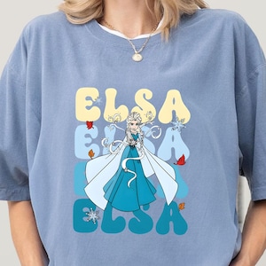 May include: A light blue t-shirt featuring a graphic of Elsa from Frozen. The word "ELSA" is stacked vertically in yellow, blue, and teal. Elsa is depicted in a blue dress and white cape, with snowflakes and leaves.
