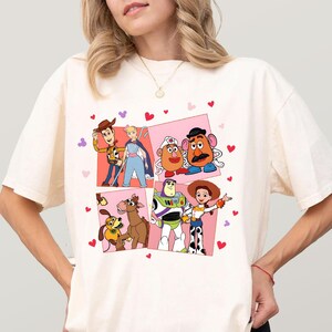 May include: Cream-coloured t-shirt featuring a colourful graphic of characters from the animated film Toy Story. The graphic includes Woody, Buzz Lightyear, Jessie, Mr. Potato Head, and others, set against a pink background with heart accents.