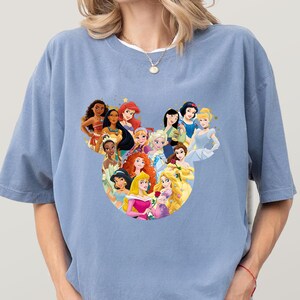 May include: A light blue t-shirt featuring a heart-shaped design composed of various cartoon princess characters. The princesses are depicted in vibrant colors, with detailed facial features and attire. The shirt has short sleeves and a relaxed fit.