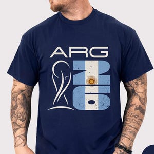 May include: Navy blue t-shirt featuring the word "ARG" above a design incorporating the Argentinian flag and a stylized trophy. The design includes the colors white, light blue, and yellow. The shirt is short-sleeved.