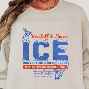 May include: A cream-coloured sweatshirt featuring a blue and red graphic that says "Kristoff & Sven's ICE Harvesting and Delivery." The design includes a snowman and reindeer illustration. The sweatshirt is long-sleeved with a crew neck.