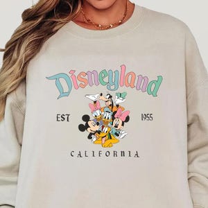 May include: A beige sweatshirt featuring the Disneyland logo with Mickey Mouse, Minnie Mouse, Donald Duck, Daisy Duck, and Goofy. The text "EST 1955 CALIFORNIA" is also printed on the sweatshirt.