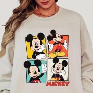 Peut inclure: Un sweat-shirt beige avec quatre panneaux repr&eacute;sentant diff&eacute;rentes images de Mickey Mouse. Chaque panneau montre Mickey dans une pose diff&eacute;rente, avec le mot "MICKEY" en lettres rouges en bas &agrave; droite.