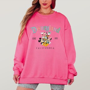 May include: A bright pink Disneyland sweatshirt. The sweatshirt features the Disneyland logo in teal and the text "EST 1955 CALIFORNIA". Below the logo are cartoon characters including Mickey Mouse, Minnie Mouse, Donald Duck, and Goofy.