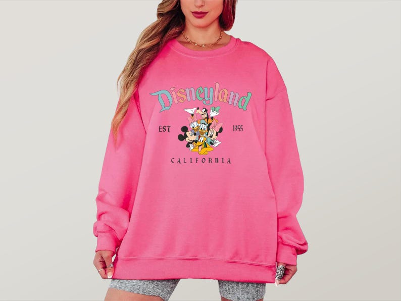 May include: A bright pink Disneyland sweatshirt. The sweatshirt features the Disneyland logo in teal and the text "EST 1955 CALIFORNIA". Below the logo are cartoon characters including Mickey Mouse, Minnie Mouse, Donald Duck, and Goofy.