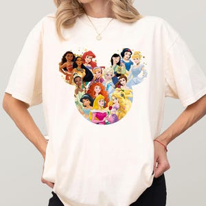 May include: Cream-colored t-shirt featuring a graphic of Disney princesses arranged in a Mickey Mouse head shape. The princesses are depicted in their signature outfits and hairstyles. The shirt is a casual, everyday wear item.
