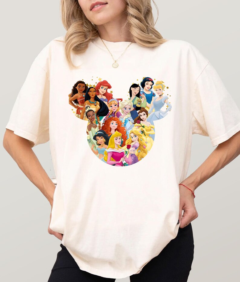 May include: Cream-colored t-shirt featuring a graphic of Disney princesses arranged in a Mickey Mouse head shape. The princesses are depicted in their signature outfits and hairstyles. The shirt is a casual, everyday wear item.