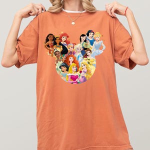 May include: An orange-colored t-shirt featuring a graphic of Disney princesses arranged in a circular design. The princesses include Snow White, Cinderella, Ariel, and others. The shirt is made of a soft material.