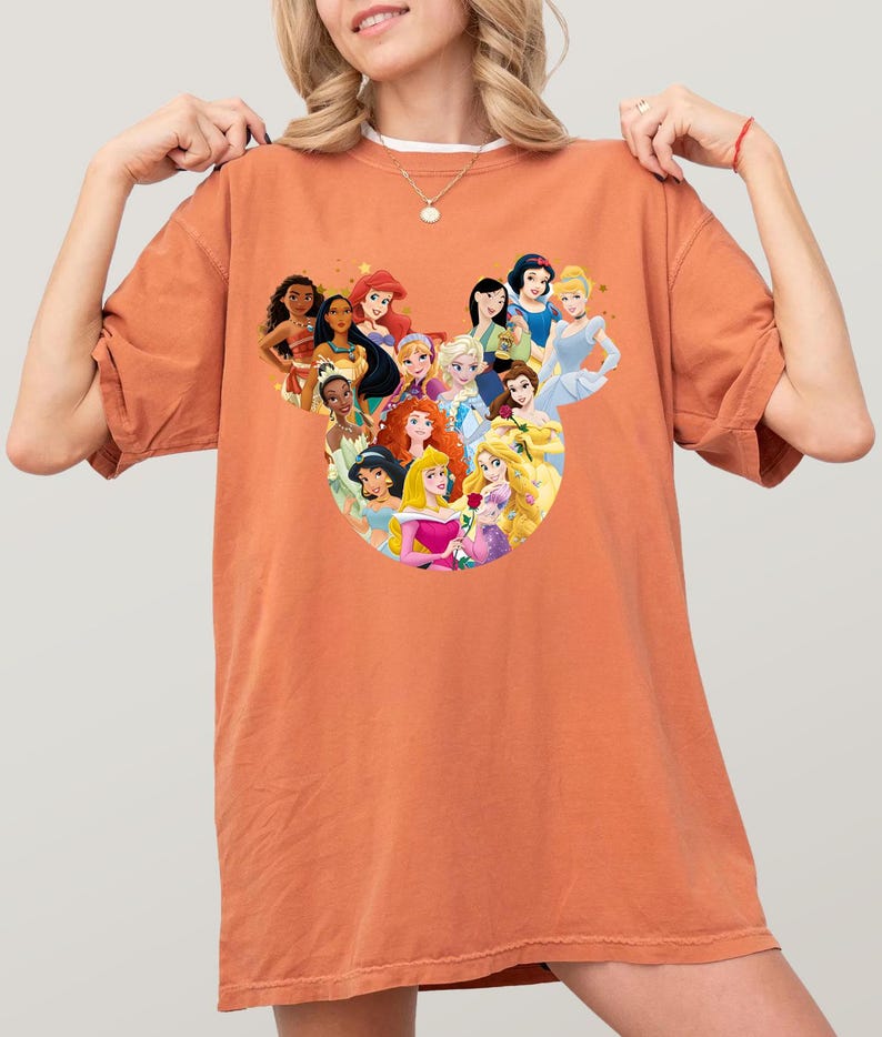 May include: An orange-colored t-shirt featuring a graphic of Disney princesses arranged in a circular design. The princesses include Snow White, Cinderella, Ariel, and others. The shirt is made of a soft material.