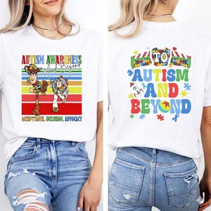 K&ouml;nnte beinhalten: Wei&szlig;es T-Shirt mit einer farbenfrohen Grafik zur F&ouml;rderung des Autismus-Bewusstseins. Auf der Vorderseite sind Woody und Buzz Lightyear mit dem Text "Autism Awareness and Beyond" und "Acceptance, Inclusion, Advocacy" abgebildet. Auf der R&uuml;ckseite steht "Autism and Beyond" mit Puzzleteilen und einem Band.