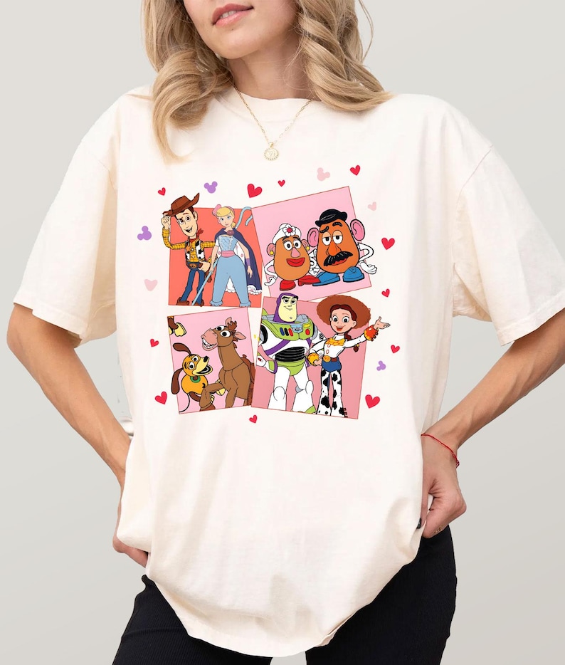 May include: Cream-coloured t-shirt featuring a colourful graphic of characters from the animated film Toy Story. The graphic includes Woody, Buzz Lightyear, Jessie, Mr. Potato Head, and others, set against a pink background with heart accents.