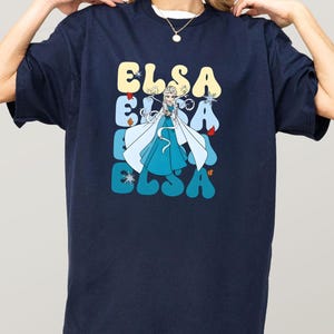 May include: Navy blue t-shirt featuring the name "ELSA" in a retro font, with a cartoon image of a character in a blue dress and white cape. The design includes snowflakes and other decorative elements.