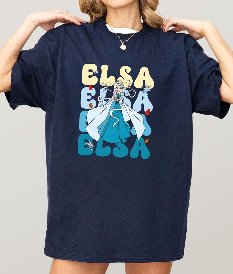 May include: Navy blue t-shirt featuring the name "ELSA" in a retro font, with a cartoon image of a character in a blue dress and white cape. The design includes snowflakes and other decorative elements.