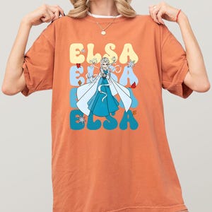 May include: Orange t-shirt featuring a graphic of Elsa from Frozen. The design includes the name "ELSA" in a retro font, with Elsa in a blue dress and white cape. The shirt has a relaxed fit.