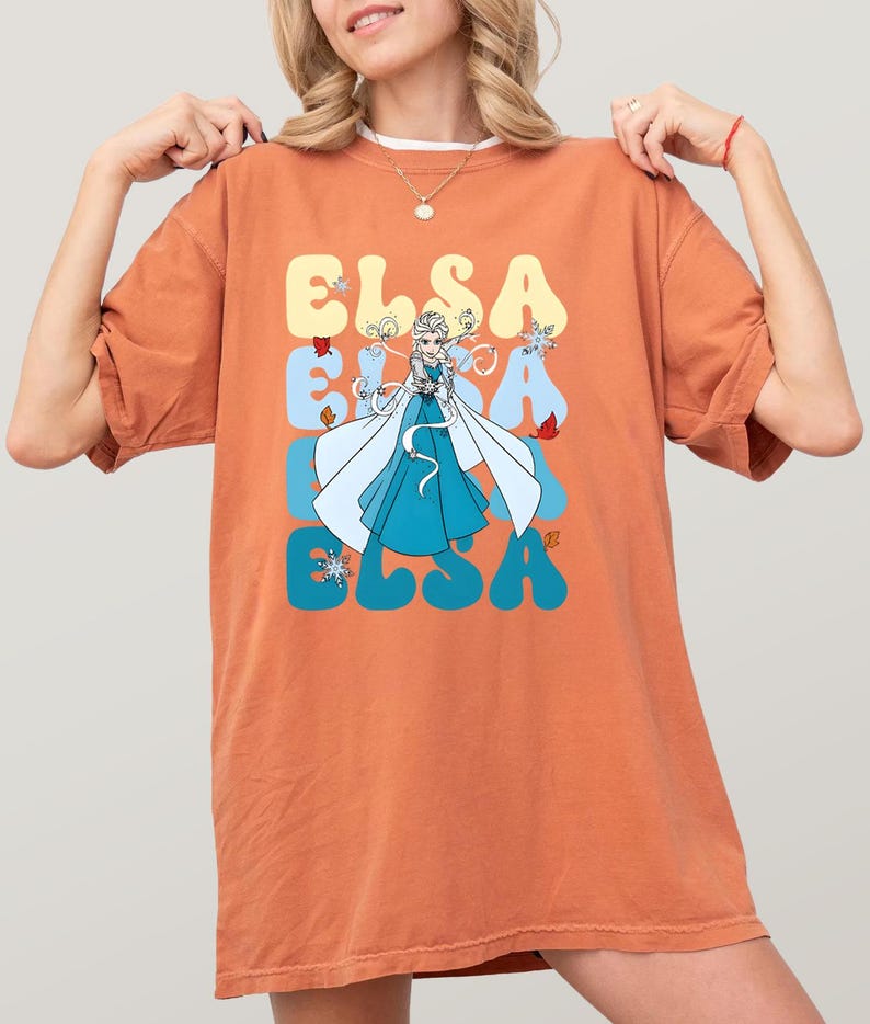 May include: Orange t-shirt featuring a graphic of Elsa from Frozen. The design includes the name "ELSA" in a retro font, with Elsa in a blue dress and white cape. The shirt has a relaxed fit.
