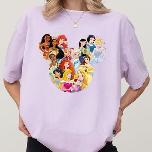 May include: A light purple t-shirt featuring a graphic of various Disney princesses arranged in a Mickey Mouse head shape. The princesses are depicted with their signature looks and colors, creating a whimsical design.