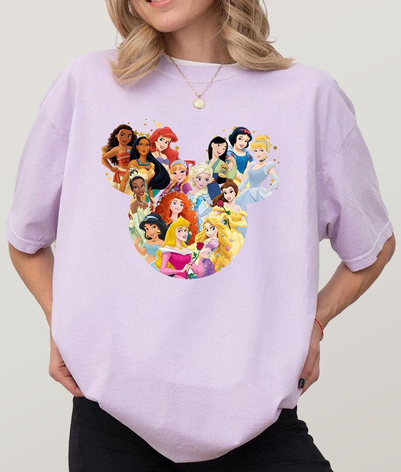 May include: A light purple t-shirt featuring a graphic of various Disney princesses arranged in a Mickey Mouse head shape. The princesses are depicted with their signature looks and colors, creating a whimsical design.