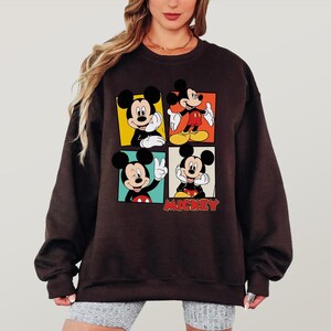 Peut inclure: Un sweat-shirt marron fonc&eacute; avec quatre panneaux repr&eacute;sentant Mickey Mouse dans diff&eacute;rentes poses. Chaque panneau a une couleur de fond diff&eacute;rente : jaune, orange, vert et rouge. Le mot "Mickey" est imprim&eacute; en rouge en bas.