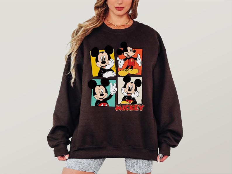 Peut inclure: Un sweat-shirt marron fonc&eacute; avec quatre panneaux repr&eacute;sentant Mickey Mouse dans diff&eacute;rentes poses. Chaque panneau a une couleur de fond diff&eacute;rente : jaune, orange, vert et rouge. Le mot "Mickey" est imprim&eacute; en rouge en bas.