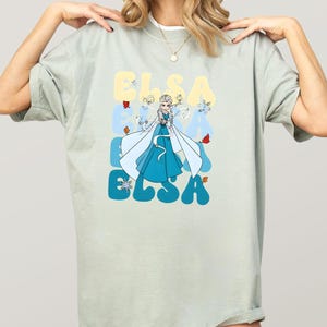 May include: A light green t-shirt featuring a graphic of Elsa from Frozen. The word "ELSA" is printed in a retro font in blue and yellow. Elsa is depicted in a blue dress with a white cape, surrounded by snowflakes and autumn leaves.