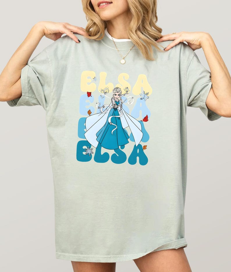 May include: A light green t-shirt featuring a graphic of Elsa from Frozen. The word "ELSA" is printed in a retro font in blue and yellow. Elsa is depicted in a blue dress with a white cape, surrounded by snowflakes and autumn leaves.
