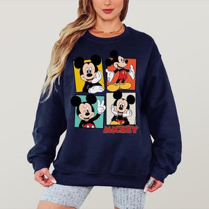 Peut inclure: Sweat-shirt bleu marine avec quatre panneaux repr&eacute;sentant Mickey Mouse dans diff&eacute;rentes poses. Chaque panneau a une couleur de fond diff&eacute;rente : jaune, orange, rouge et noir. Le mot "Mickey" est imprim&eacute; en rouge en bas.