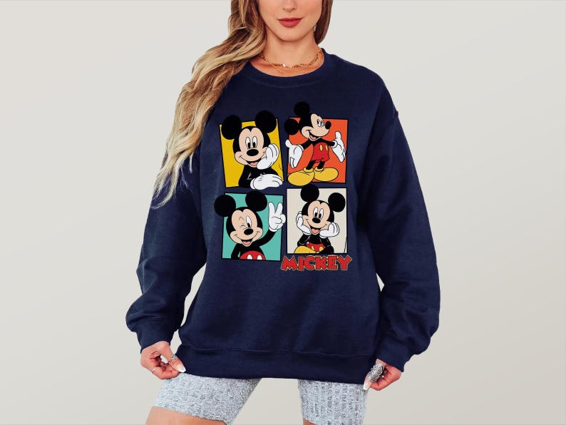 Peut inclure: Sweat-shirt bleu marine avec quatre panneaux repr&eacute;sentant Mickey Mouse dans diff&eacute;rentes poses. Chaque panneau a une couleur de fond diff&eacute;rente : jaune, orange, rouge et noir. Le mot "Mickey" est imprim&eacute; en rouge en bas.