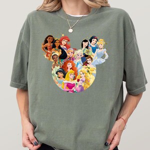 May include: Sage green t-shirt featuring a graphic of various animated princesses arranged in a Mickey Mouse head shape. The princesses are depicted with vibrant colors and detailed designs, creating a whimsical and eye-catching design.
