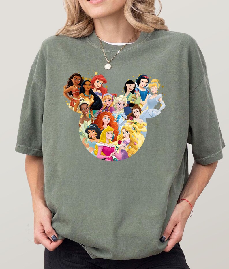 May include: Sage green t-shirt featuring a graphic of various animated princesses arranged in a Mickey Mouse head shape. The princesses are depicted with vibrant colors and detailed designs, creating a whimsical and eye-catching design.