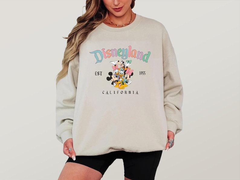 May include: A beige sweatshirt featuring the Disneyland logo with Mickey Mouse, Minnie Mouse, Donald Duck, Daisy Duck, and Goofy. The text "EST 1955 CALIFORNIA" is also printed on the sweatshirt.