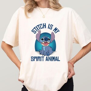 Puede incluir: Camiseta color crema con una ilustraci&oacute;n de Stitch, el personaje azul sonriendo. El texto "STITCH IS MY SPIRIT ANIMAL" en azul est&aacute; escrito encima y debajo de la imagen.