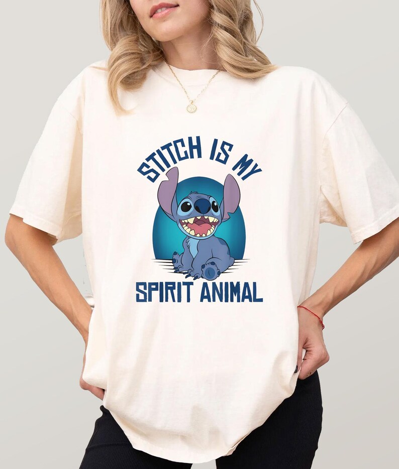 Puede incluir: Camiseta color crema con una ilustraci&oacute;n de Stitch, el personaje azul sonriendo. El texto "STITCH IS MY SPIRIT ANIMAL" en azul est&aacute; escrito encima y debajo de la imagen.