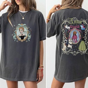 May include: Dark gray t-shirt featuring a cartoon image of a character in a black dress and a teal bow. The back of the shirt has the name "Anna" and a framed image of the character in a pink dress, with other cartoon characters.