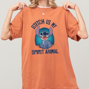Puede incluir: Camiseta naranja con el personaje Stitch de Lilo & Stitch. El gr&aacute;fico azul muestra a Stitch con el texto "Stitch is my spirit animal" en azul. La camiseta es de color naranja liso.