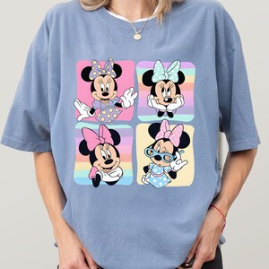 Może przedstawiać: Jasnoniebieska koszulka z kolorową grafiką Myszki Minnie w czterech r&oacute;żnych pozach. Każda poza jest umieszczona na tle pastelowej tęczy. Myszka Minnie nosi kokardy, sukienki i okulary.