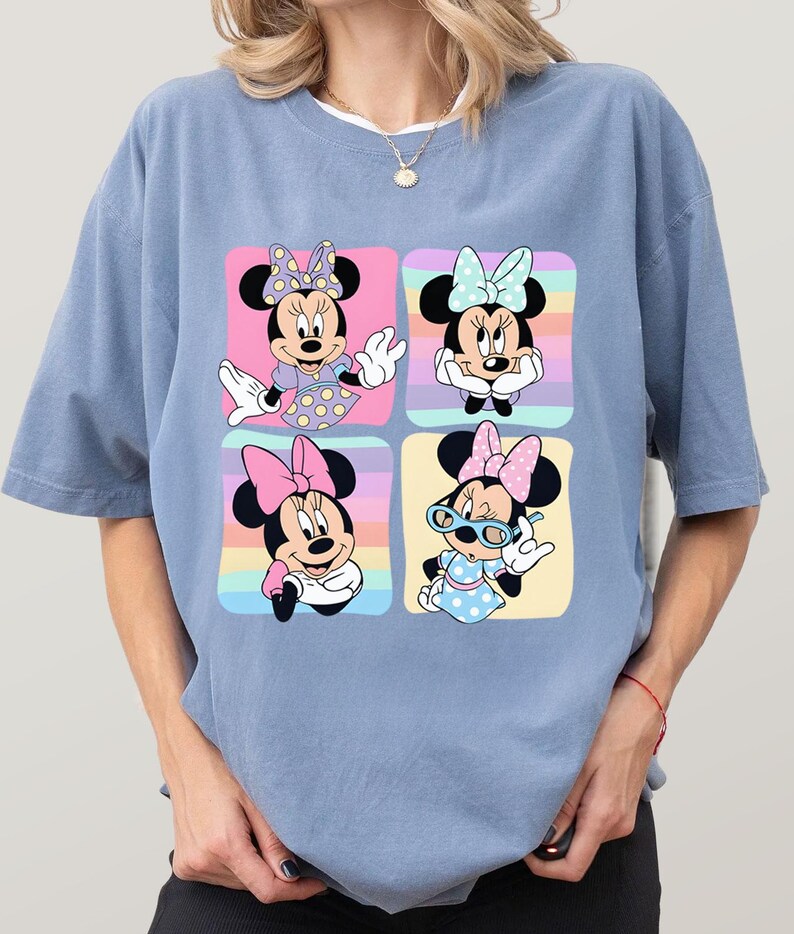Może przedstawiać: Jasnoniebieska koszulka z kolorową grafiką Myszki Minnie w czterech r&oacute;żnych pozach. Każda poza jest umieszczona na tle pastelowej tęczy. Myszka Minnie nosi kokardy, sukienki i okulary.