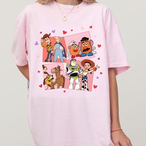 May include: Pale pink t-shirt with a vibrant graphic of Toy Story characters. The design showcases Woody, Buzz Lightyear, Jessie, and others, set against a backdrop of pink squares and heart shapes.