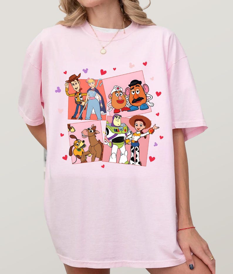 May include: Pale pink t-shirt with a vibrant graphic of Toy Story characters. The design showcases Woody, Buzz Lightyear, Jessie, and others, set against a backdrop of pink squares and heart shapes.