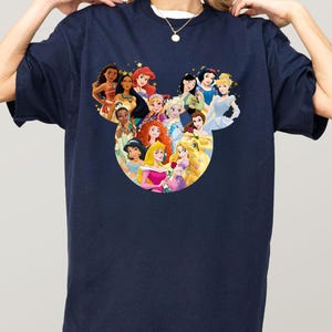 May include: Navy blue t-shirt featuring a graphic of Disney princesses arranged in a Mickey Mouse head shape. The princesses are depicted in their signature outfits and surrounded by gold stars. The shirt is a casual, oversized style.