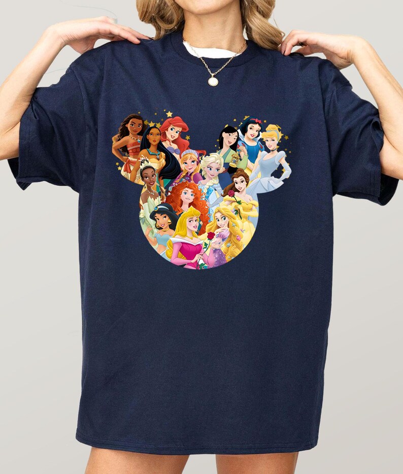 May include: Navy blue t-shirt featuring a graphic of Disney princesses arranged in a Mickey Mouse head shape. The princesses are depicted in their signature outfits and surrounded by gold stars. The shirt is a casual, oversized style.
