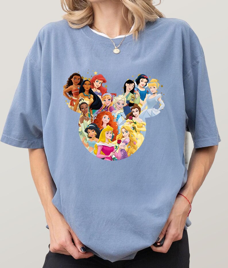 May include: A light blue t-shirt featuring a heart-shaped design composed of various cartoon princess characters. The princesses are depicted in vibrant colors, with detailed facial features and attire. The shirt has short sleeves and a relaxed fit.